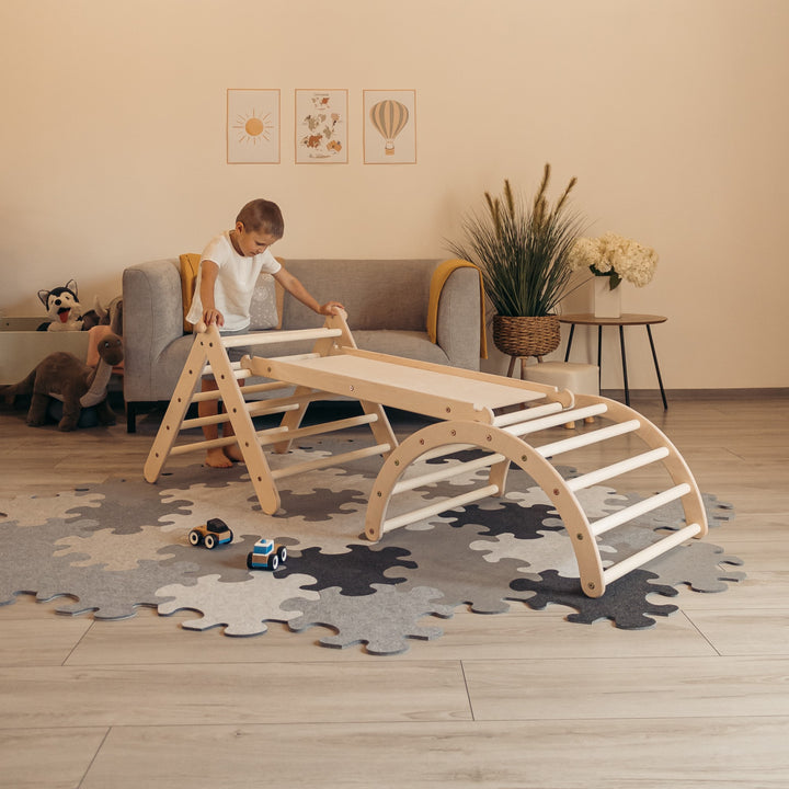 A child playing with a wooden playset consisting of an adjustable triangle, a chicken ladder slide, and a arched structure, placed in a room with a puzzle mat on the floor.