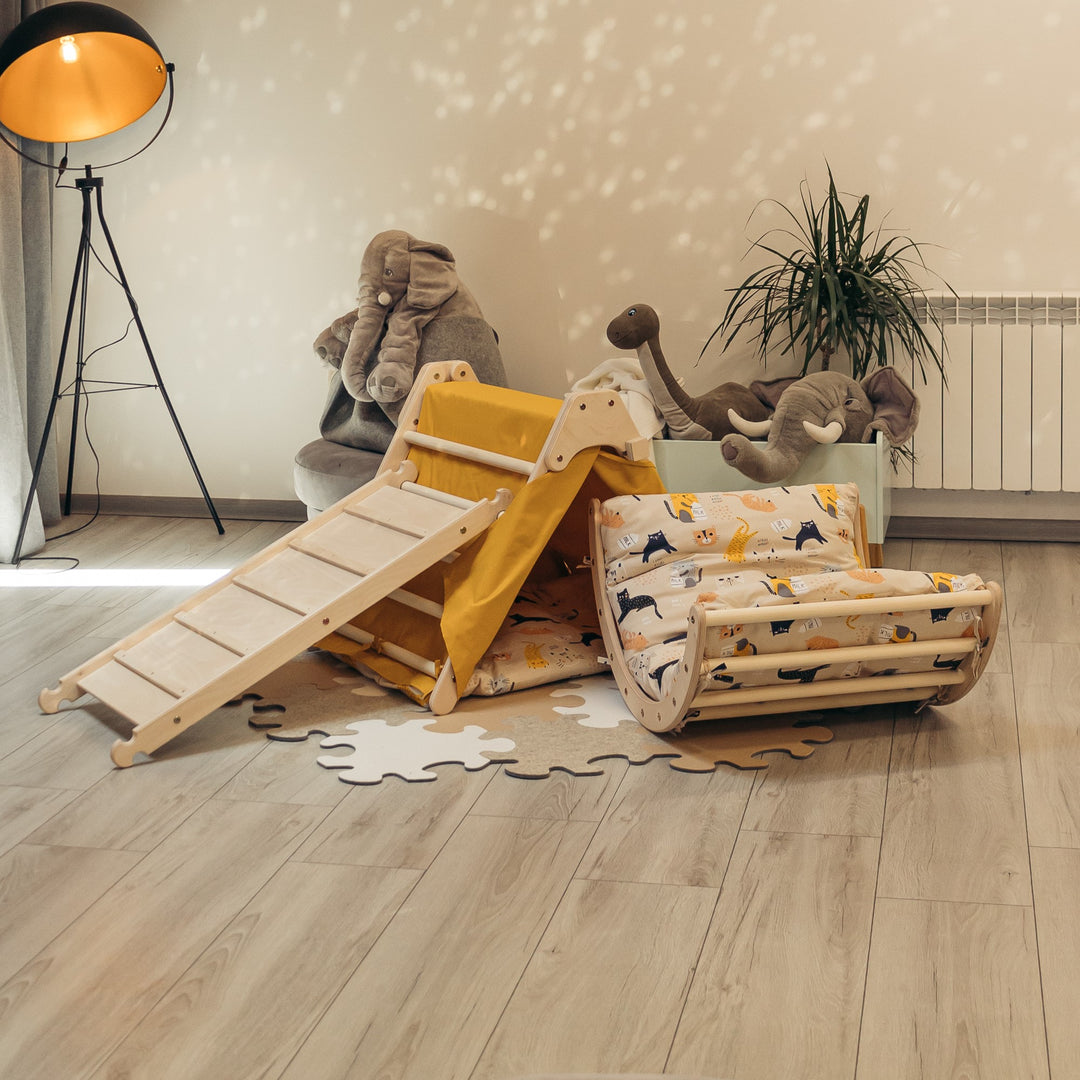 Children's play set including a fold-up triangle, a sliding board, and a small arch with mustard-colored accents. The set is arranged on a wooden floor with soft toys around.