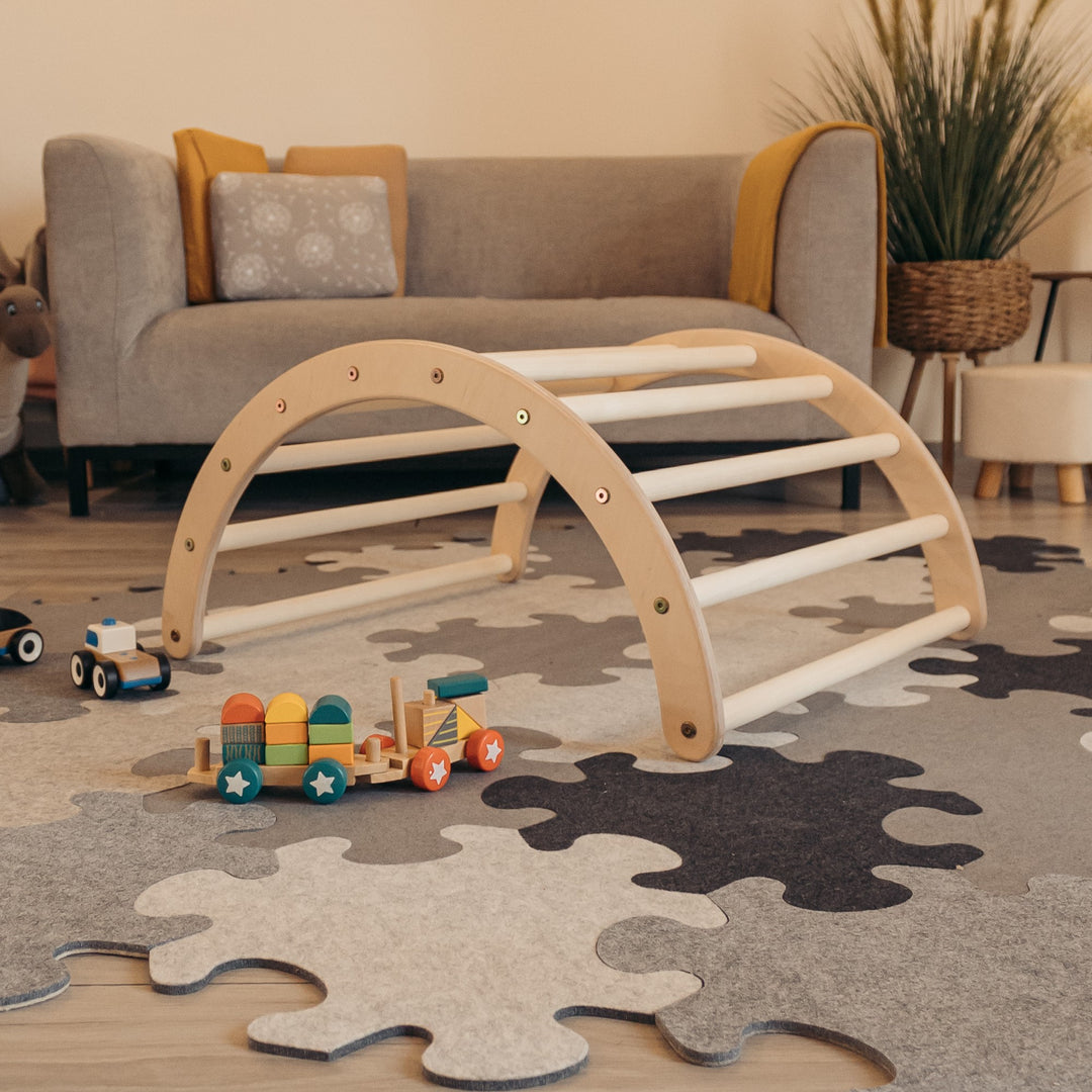 Children's wooden climbing arch toy placed on the floor in a room, with a couch and a toy train set in the background.