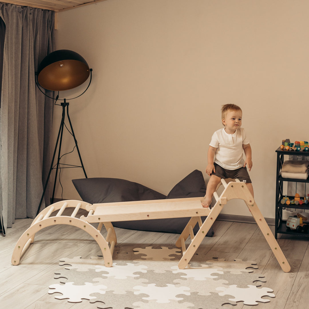 A young child playing on a wooden fold-up triangle with a chicken ladder slide and an arched structure, placed in a room with toys around. Climb up toys